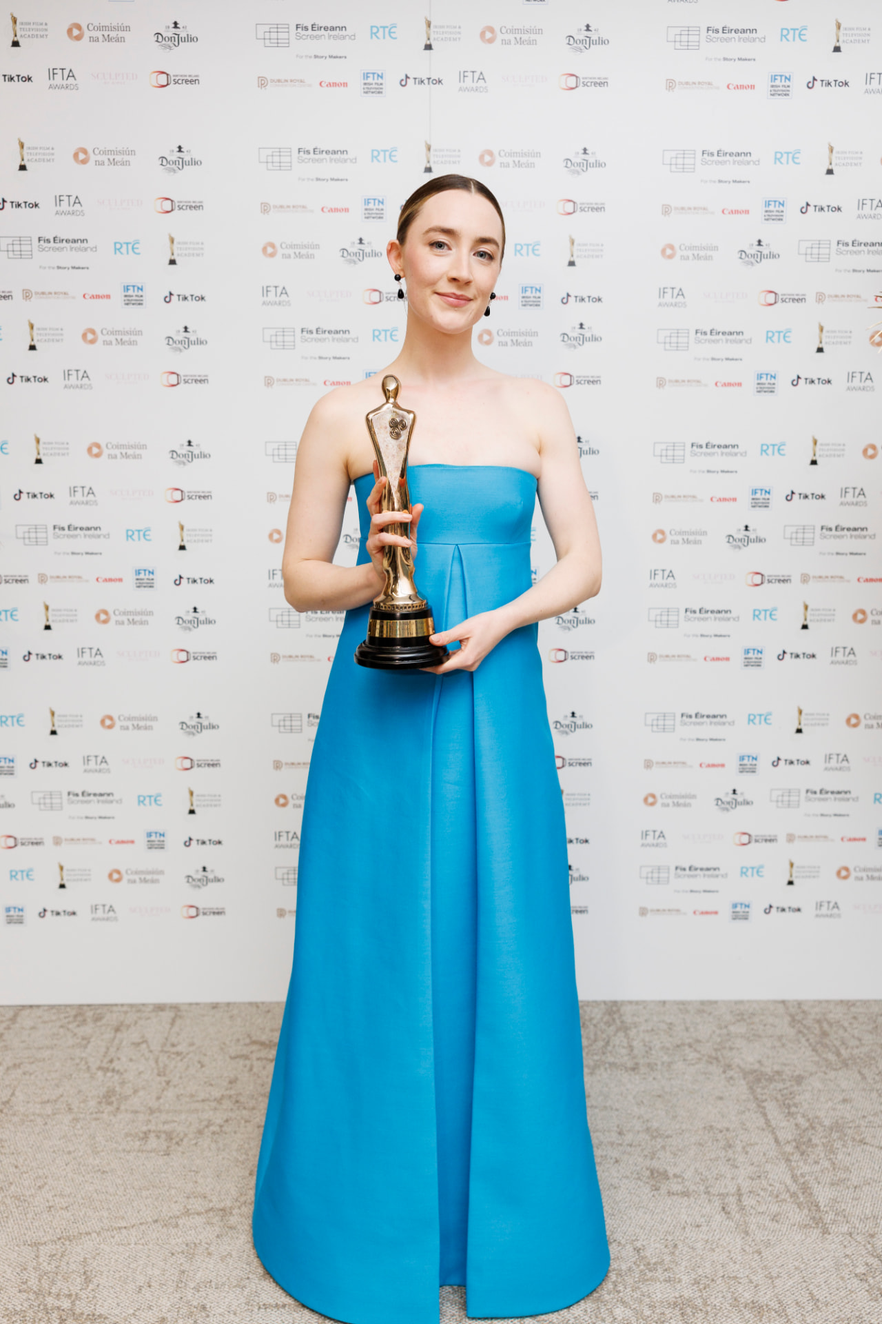 Guest in a turquoise floor-length gown holding an award in front of the IFTA press backdrop