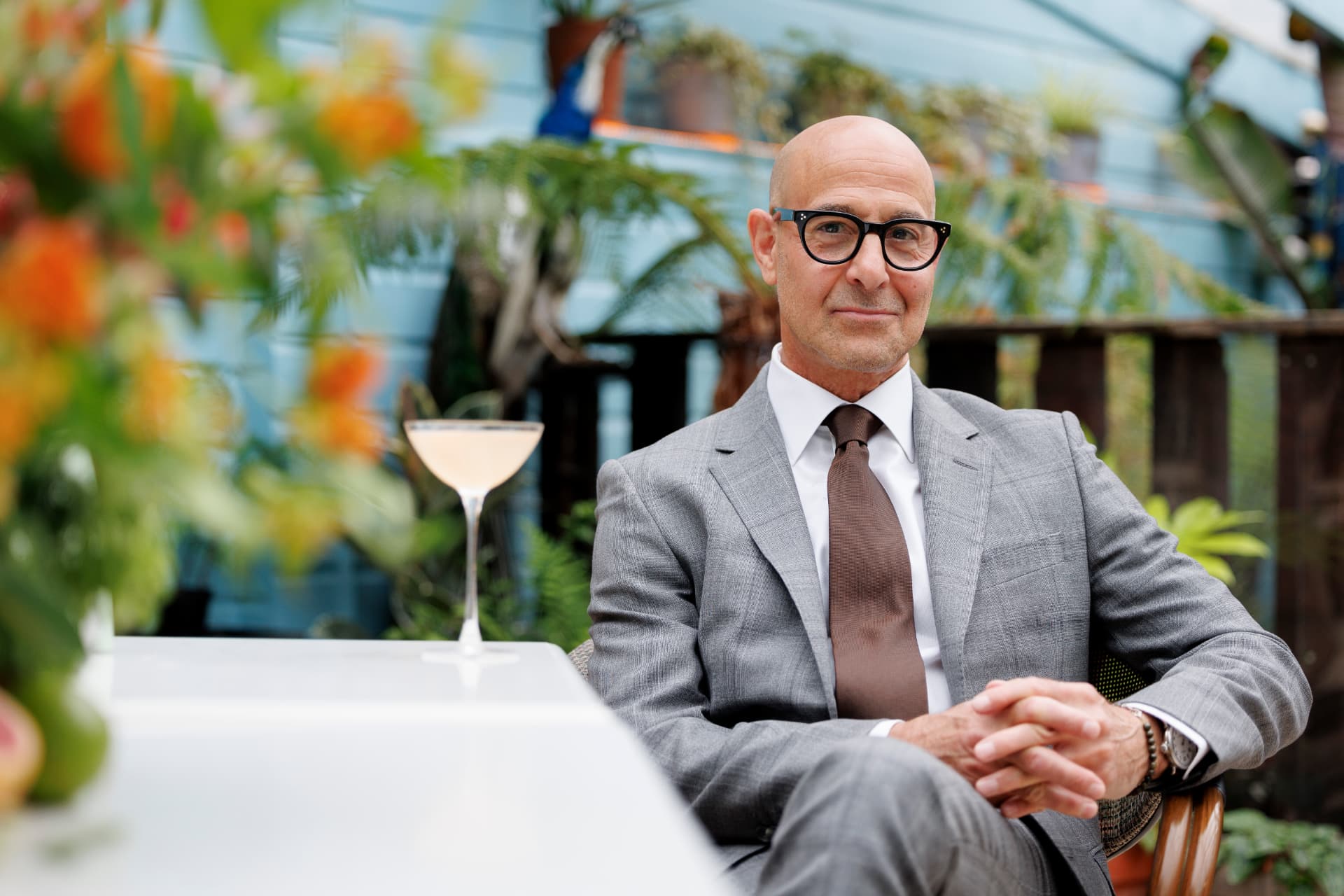 Stanley Tucci seated outdoors in a grey suit and glasses, holding a cocktail for a Tanqueray portrait